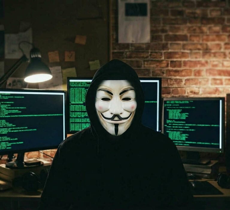 Hacker wearing a Guy Fawkes mask working on multiple computer screens with code, symbolising cyber s.